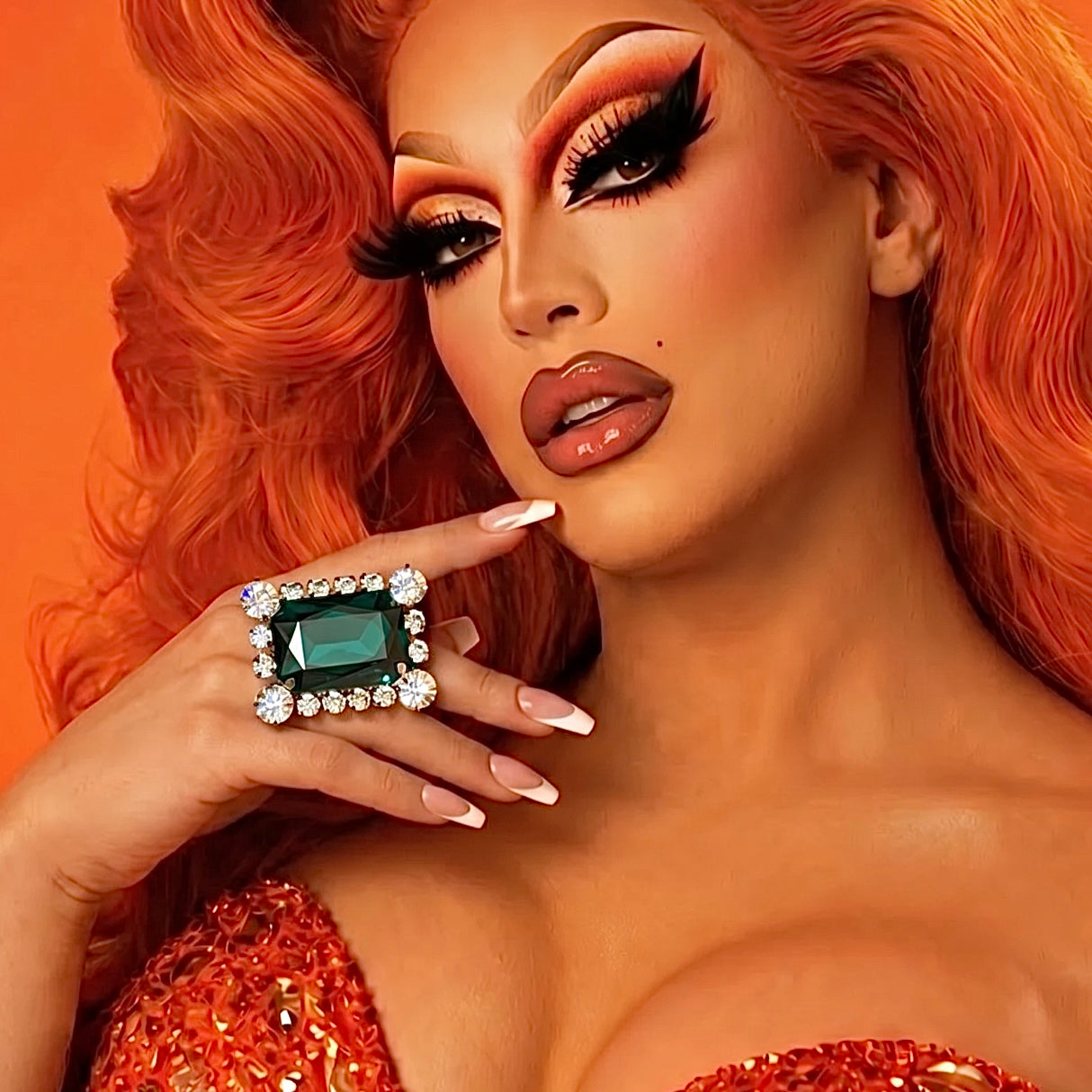 Woman with vibrant makeup and red hair holding a green gemstone ring against an orange background