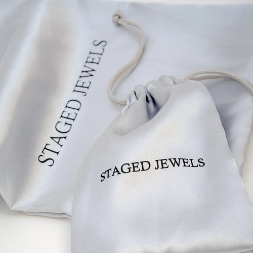 White drawstring bag with 'STAGED JEWELS' branding on a light gray background