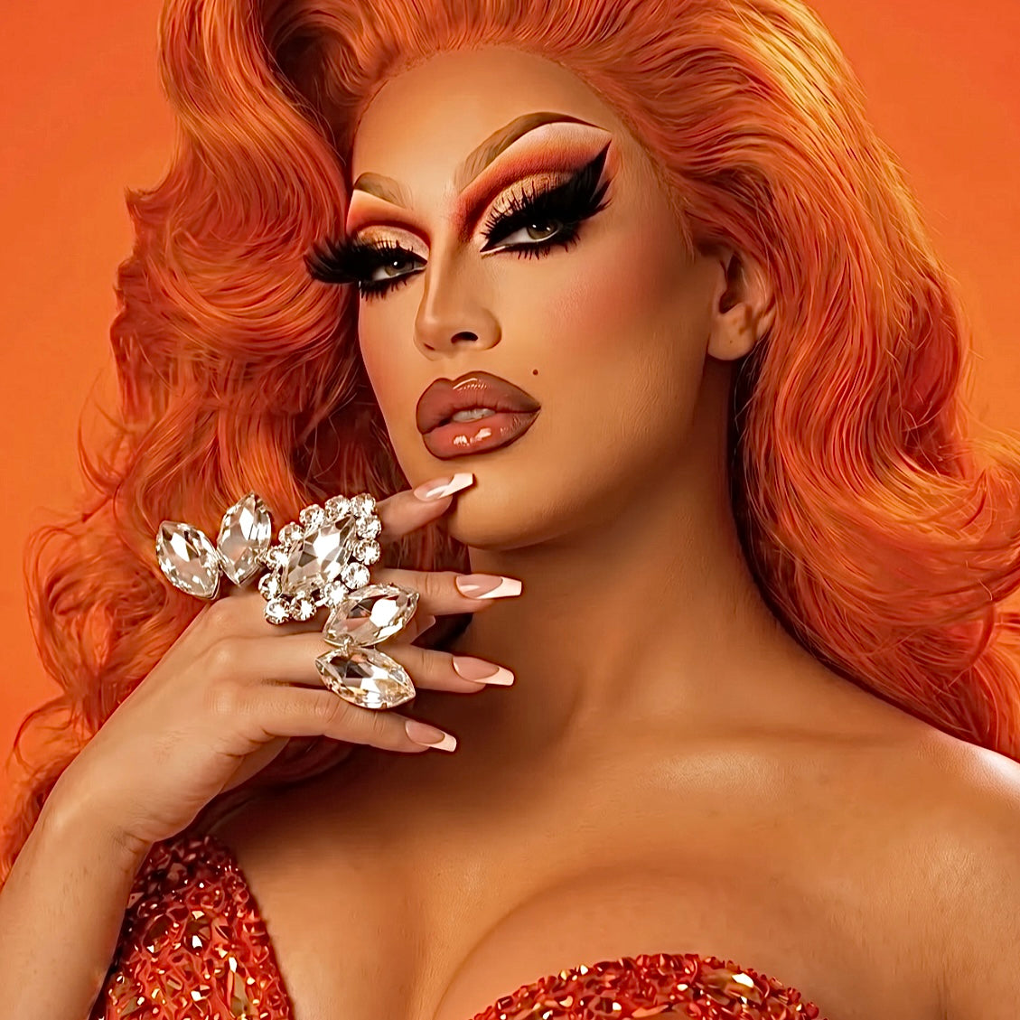 Person with vibrant orange hair and makeup, wearing a sparkling top against an orange background